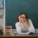 Professora de óculos com as mãos na cabeça demonstrando cansaço e sobrecarga ao elaborar relatórios de alunos em mesa de sala de aula com ábaco e quadro verde ao fundo