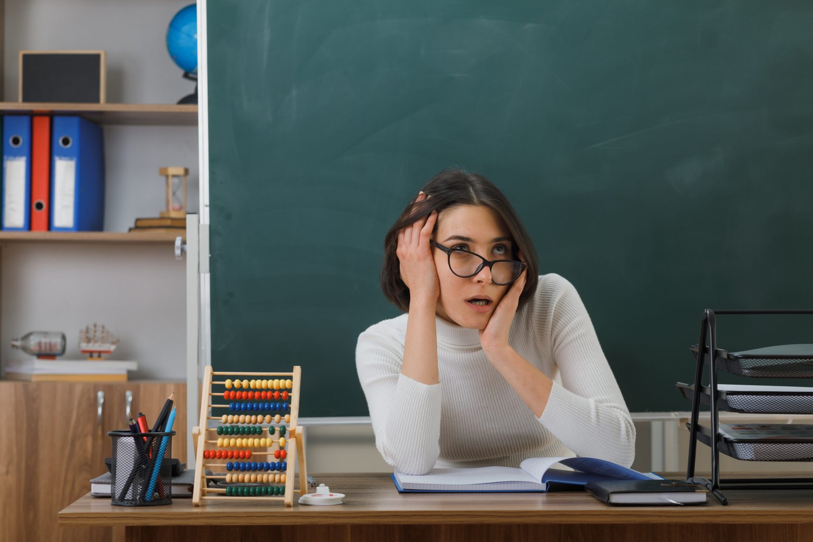 Professora de óculos com as mãos na cabeça demonstrando cansaço e sobrecarga ao elaborar relatórios de alunos em mesa de sala de aula com ábaco e quadro verde ao fundo